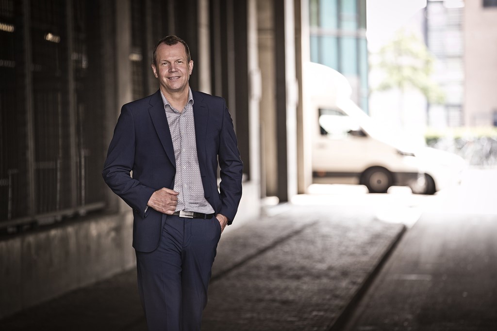 Mads Moesgaard stands smiling outside a large office building. Wearing dark suit trousers and a wildly patterned shirt, he discreetly straightens his shirt sleeve.