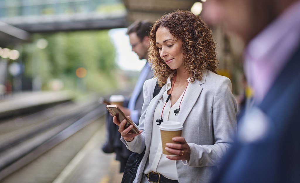 Woman standing at train station with a take away coffee in one hand, mobile phone in other. She has curly hair and looks relaxed in bright office clothes.