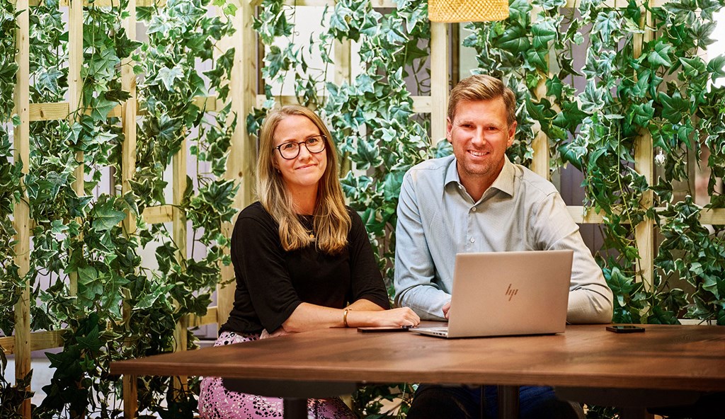 Two colleagues are sitting by a computer and smiling at the camera. Behind them they have a wall of climbing plants.