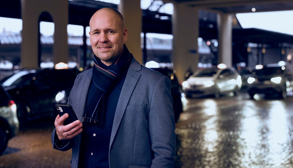 Lars har en mobiltelefon i hånden og står ved siden av en vei hvor mange drosjer kjører forbi. Han har dress og et tykt skjerf.